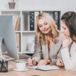 smiling female business mentor pointing at notebook and working with young colleague in office