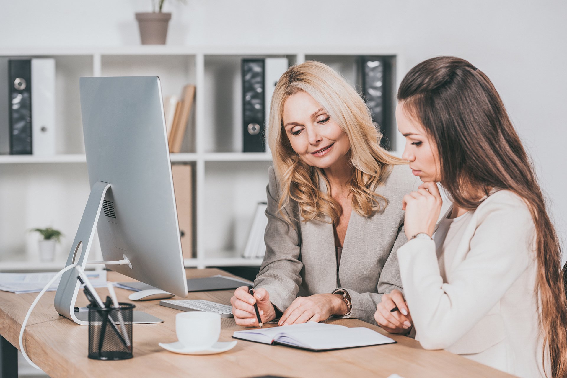 smiling female business mentor pointing at notebook and working with young colleague in office